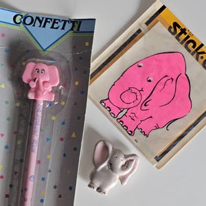 May include: A collection of pink elephant-themed stationery and stickers. Includes a pink elephant pencil, a pink elephant sticker, and a small ceramic elephant figurine. The pencil is packaged in a "Confetti" themed package.