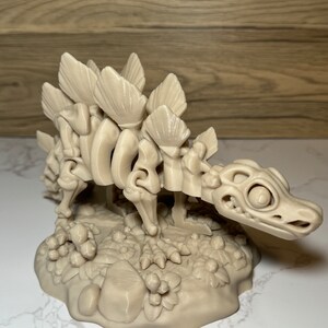 Pu&ograve; includere: Un modello scheletrico di dinosauro stampato in 3D di colore beige, con una schiena spinosa, in piedi su una base con una superficie testurizzata.