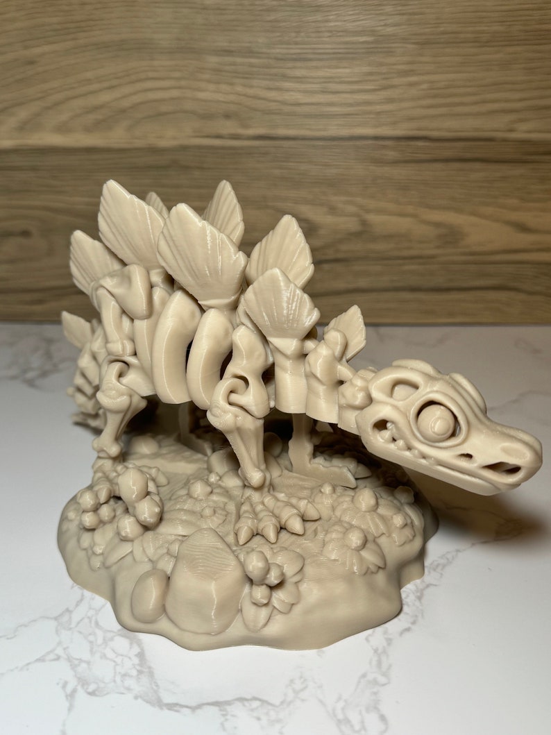 Pu&ograve; includere: Un modello scheletrico di dinosauro stampato in 3D di colore beige, con una schiena spinosa, in piedi su una base con una superficie testurizzata.