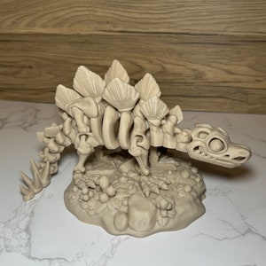 Pu&ograve; includere: Un modello di dinosauro scheletrico di colore beige stampato in 3D con una coda spinosa e una fila di piastre lungo la schiena. Il dinosauro &egrave; in piedi su una base con una superficie testurizzata.