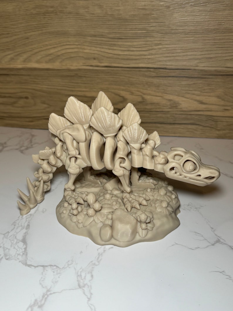 Pu&ograve; includere: Un modello di dinosauro scheletrico di colore beige stampato in 3D con una coda spinosa e una fila di piastre lungo la schiena. Il dinosauro &egrave; in piedi su una base con una superficie testurizzata.