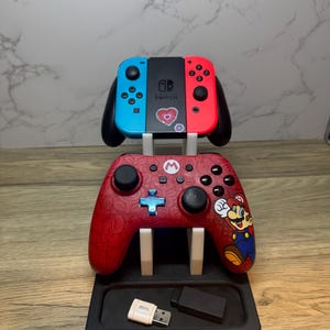 Può includere: Un controller Nintendo Switch rosso e nero e i Joy-Con sono esposti su un supporto nero. Il controller presenta una grafica di Mario. Un adattatore USB e un dongle nero sono nella base del supporto. I Joy-Con sono blu e rossi.