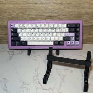 Puede incluir: Un teclado mecánico morado y blanco con teclas negras y un soporte de teclado negro. El teclado tiene una fila de teclas de función y una fila de teclas numéricas. El soporte de teclado está hecho de metal y tiene un diseño simple.