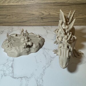 Pu&ograve; includere: Una statuetta di drago grigia stampata in 3D con un design scheletrico dettagliato. Il drago &egrave; in piedi su una superficie di marmo bianco.