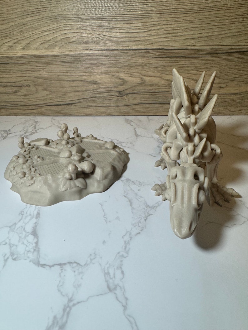 Pu&ograve; includere: Una statuetta di drago grigia stampata in 3D con un design scheletrico dettagliato. Il drago &egrave; in piedi su una superficie di marmo bianco.