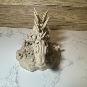 Pu&ograve; includere: Una statuetta di drago grigia stampata in 3D con una schiena spinosa e squame dettagliate. Il drago &egrave; in posa su una base rocciosa con piccole piante.