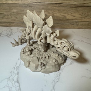 Pu&ograve; includere: Un modello stampato in 3D di uno scheletro di Stegosauro, posto su una base con una texture rocciosa e frondosa. Il modello &egrave; di colore beige e presenta dettagli intricati delle ossa del dinosauro.