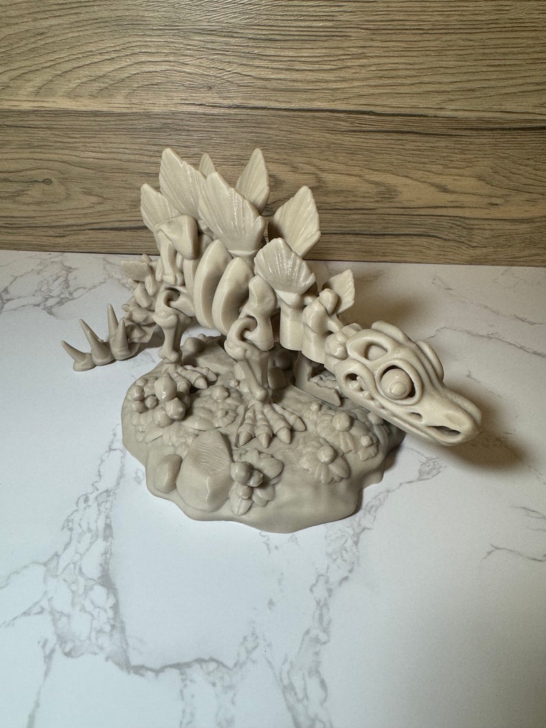 Pu&ograve; includere: Un modello stampato in 3D di uno scheletro di Stegosauro, posto su una base con una texture rocciosa e frondosa. Il modello &egrave; di colore beige e presenta dettagli intricati delle ossa del dinosauro.