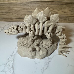 Pu&ograve; includere: Un modello di scheletro di dinosauro in plastica beige con una coda spinosa e una fila di piastre lungo la schiena. Il modello &egrave; posto su una base con una superficie testurizzata.