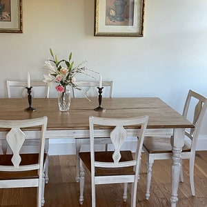 May include: A white farmhouse style dining table with a distressed wood top and white painted legs. The table is set with six chairs, two of which are visible in the image. The chairs have a white painted finish and a brown wood seat. There are white candlesticks and a vase of white and pink flowers on the table.