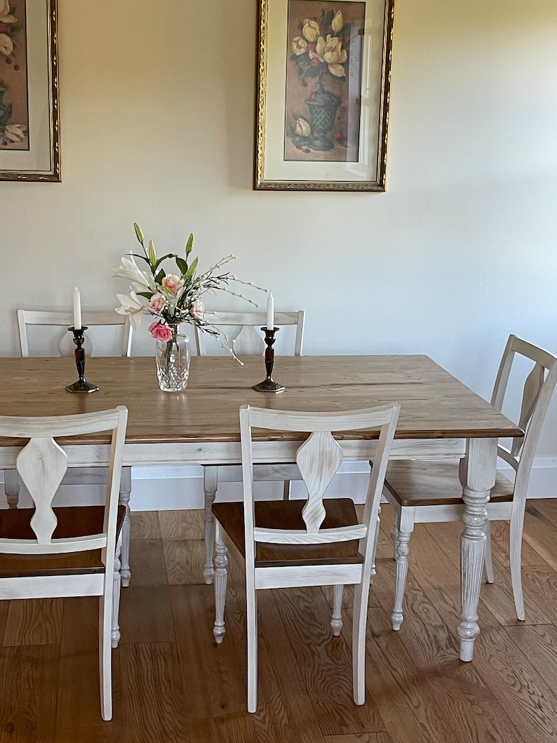 May include: A white farmhouse style dining table with a distressed wood top and white painted legs. The table is set with six chairs, two of which are visible in the image. The chairs have a white painted finish and a brown wood seat. There are white candlesticks and a vase of white and pink flowers on the table.