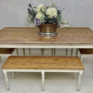 May include: A white farmhouse style dining table with a wood top and turned legs. The table is set with four chairs and a bench. A vase of white and purple flowers sits in the centre of the table.
