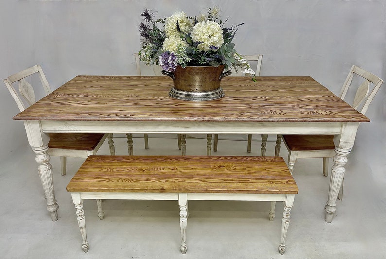 May include: A white farmhouse style dining table with a wood top and turned legs. The table is set with four chairs and a bench. A vase of white and purple flowers sits in the centre of the table.