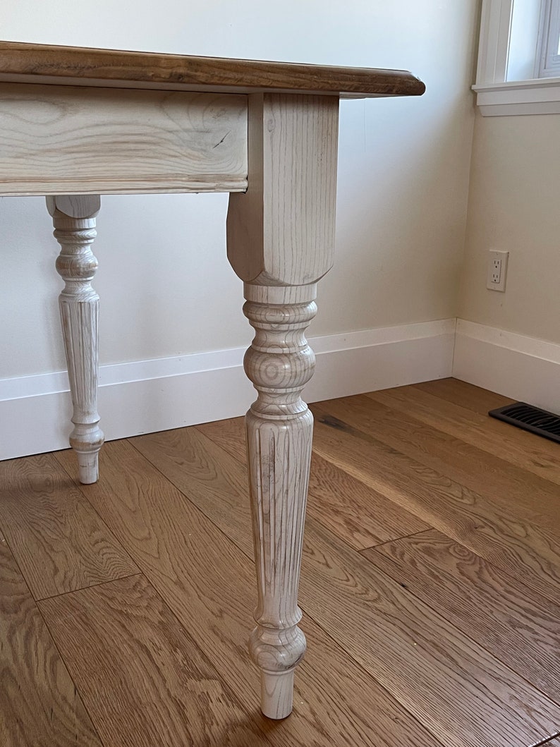 May include: White painted wooden table with a natural wood top and turned legs. The table has a simple, farmhouse style.