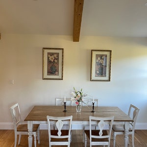 May include: A white farmhouse dining table with six chairs. The table has a distressed white finish and a wood top. The chairs are also white with a distressed finish. There are two framed pictures on the wall behind the table. The pictures are of flowers in vases.