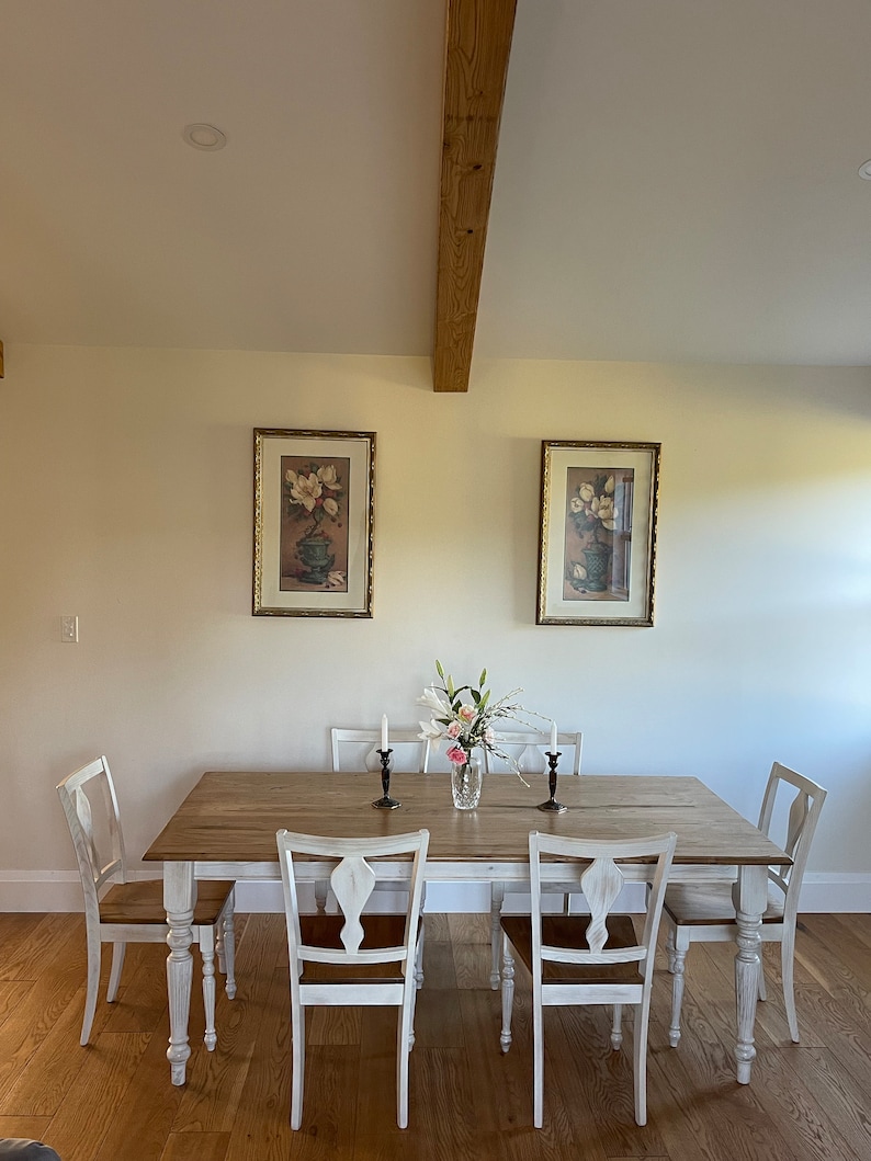 May include: A white farmhouse dining table with six chairs. The table has a distressed white finish and a wood top. The chairs are also white with a distressed finish. There are two framed pictures on the wall behind the table. The pictures are of flowers in vases.