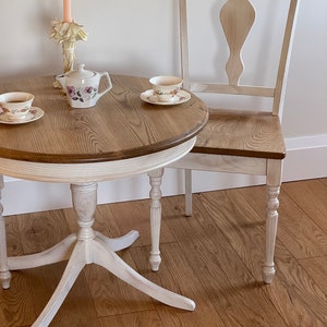 Peut inclure: Une table ronde blanche avec un plateau en bois et une chaise blanche avec un siège en bois. La table a un piétement en colonne avec des pieds tournés. La chaise a un dossier à fuseaux et des pieds tournés. Il y a deux tasses à thé et soucoupes sur la table, ainsi qu'une théière et un bougeoir.