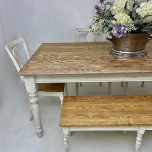May include: White farmhouse style dining table with a wood top and white painted legs. The table is set with a white chair and a bench with a wood top and white painted legs. A vase of white and purple flowers sits on the table.