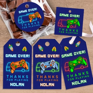 Peut inclure: Etiquettes de faveur bleues et noires avec un graphisme de manette de jeu vidéo vert néon, orange et bleu. Le texte sur les étiquettes indique "Game Over ! Merci d’avoir joué Nolan".