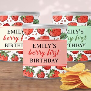 Może przedstawiać: Trzy srebrne puszki z etykietami z napisem "Emily's berry first birthday" i ozdobione czerwonymi truskawkami na białym tle. Puszki stoją na drewnianej powierzchni z kupką chipsów ziemniaczanych z przodu.