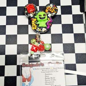 May include: A black badge reel with a green character design, red dice, and colorful accents. The reel has a clear plastic holder with a business card that reads "Salgado's" and lists various crafts. A charm of beads and red roses hangs below.