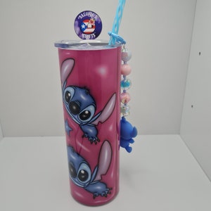 Puede incluir: Un vaso de acero inoxidable rosa con un dise&ntilde;o de personaje de dibujos animados azul y blanco. El vaso tiene una pajita y un amuleto unido a la tapa. El amuleto es un personaje de dibujos animados azul con un lazo rosa. El vaso tiene el texto "Salgado's Crafts" en la tapa.