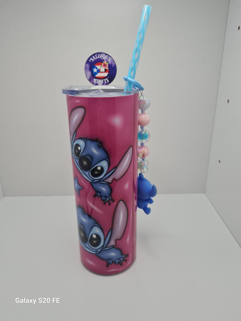Puede incluir: Un vaso de acero inoxidable rosa con un dise&ntilde;o de personaje de dibujos animados azul y blanco. El vaso tiene una pajita y un amuleto unido a la tapa. El amuleto es un personaje de dibujos animados azul con un lazo rosa. El vaso tiene el texto "Salgado's Crafts" en la tapa.
