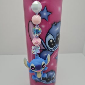 Puede incluir: Un vaso de acero inoxidable rosa con un personaje de dibujos animados azul en &eacute;l. El vaso tiene un encanto de cuentas azules y blancas adjunto.