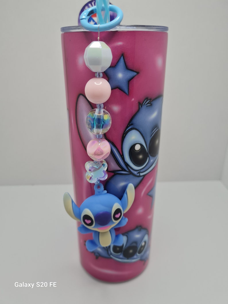 Puede incluir: Un vaso de acero inoxidable rosa con un personaje de dibujos animados azul en &eacute;l. El vaso tiene un encanto de cuentas azules y blancas adjunto.
