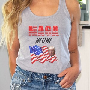 May include: A gray tank top with the text "MAGA MOM" in red and white letters. The text is layered over a waving American flag with a portrait of Donald Trump in the upper right corner.