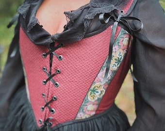May include: A red and black corset-style top with a lace-trimmed neckline and black ribbon ties. The bodice features a textured pattern and floral embroidered accents. The sleeves are sheer black fabric, and the overall style is reminiscent of historical fashion.