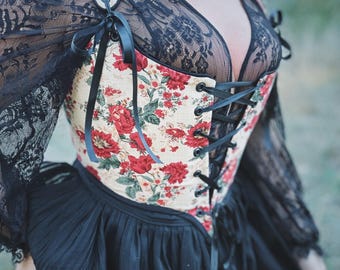 May include: A white corset with a floral print featuring red roses and green leaves. The corset has black ribbon ties and is worn over a black lace top.