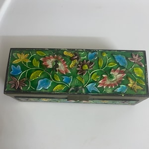 May include: A rectangular metal box featuring a vibrant green enamel background decorated with colorful floral patterns in shades of red, yellow, and blue. The lid displays intricate raised metal outlines of flowers and leaves, with a small latch on the front.