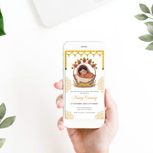 May include: A mobile phone displaying a digital invitation for a baby naming ceremony. The invitation features a golden design with a picture of a baby in a basket. The text on the invitation reads "You are invited to Amit and Amand's daughter's Naming Ceremony 17 October, 20XX | at 9:00 AM at 123 Anywhere St. Any City, ST"