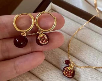 Può includere: Set di gioielli color oro con orecchini a cerchio e collana. Gli orecchini hanno un design a cerchio rotondo con un ciondolo a forma di melograno rosso scuro. La collana ha una catena delicata con un ciondolo a forma di melograno abbinato. I ciondoli a forma di melograno hanno un design dettagliato.