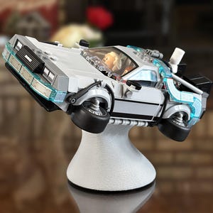 May include: A silver and black DeLorean time machine model, with blue accents, is displayed on a white stand. The model has black wheels and a detailed interior. The stand is white and has a rounded base.