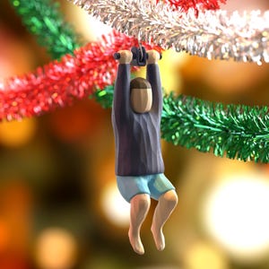 Könnte beinhalten: Ein skurriler Weihnachtsschmuck, der eine Figur zeigt, die an einer silbernen, roten und grünen Girlande hängt. Die Figur trägt ein blaues Hemd, blaue Shorts und eine beige Kappe. Der Hintergrund ist verschwommen mit warmen Farbtönen.
