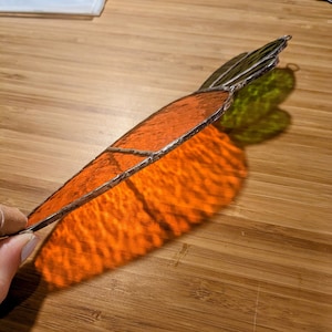 Pode incluir: Cenoura de vitral com corpo laranja e folhas verdes. A cenoura é contornada em prata e projeta uma sombra laranja vibrante. Uma peça decorativa artesanal.