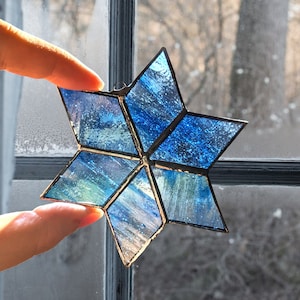 Puede incluir: Una estrella de vidrio coloreado azul y verde con un acabado metálico. La estrella está hecha de múltiples piezas de vidrio que se unen con plomo. La estrella tiene un colgador en la parte superior.