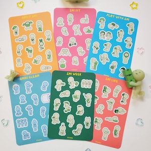 May include: A collection of colorful sticker sheets featuring whimsical illustrations of stick figures in various poses. Each sheet is a different color and has text such as "Daily Shi", "Sai Fit", and "Play With Sai". Decorative figurines and star-shaped embellishments are scattered around.