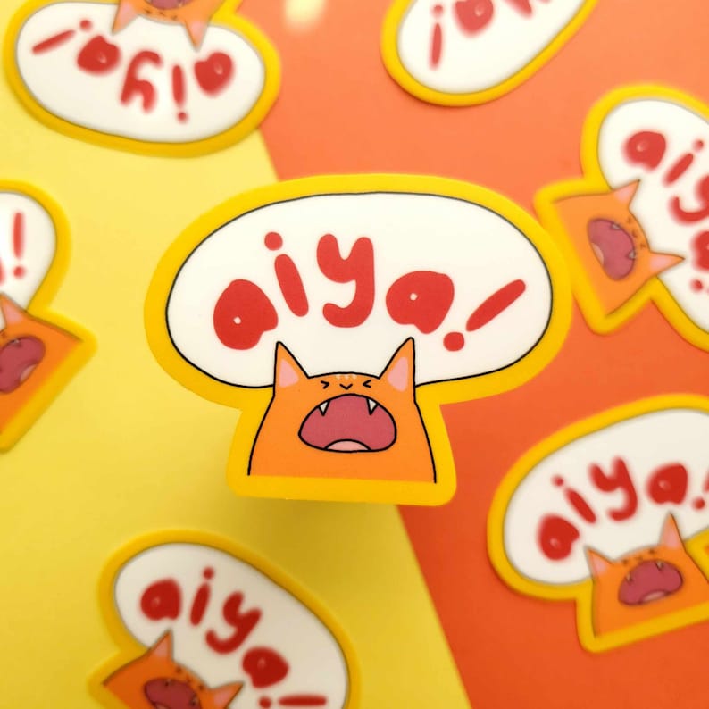 May include: A set of orange and yellow stickers featuring a cartoon cat with a speech bubble saying "aiya!" The cat is yelling with its mouth open and its eyes wide.