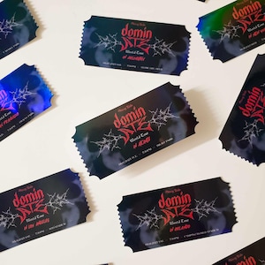 May include: A set of holographic concert ticket stubs for the "Domin Date" world tour. The tickets are black with red text and feature a graphic of a spider web. The text on the tickets reads "Domin Date World Tour" and lists the city and date of each concert.