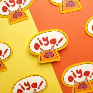 May include: A set of five orange and yellow stickers featuring a cartoon cat with a speech bubble that says "aiya!" The cat is red and has a wide open mouth. The stickers are on a yellow and orange background.