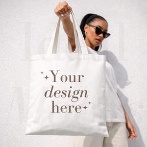Op de afbeelding: Een witte canvas draagtas met de bruine tekst Your design here, vergezeld door kleine vierpuntige sterpictogrammen. De tas heeft twee stevige schouderbanden en een rechthoekige vorm, wat een oppervlak biedt voor het plaatsen van ontwerpen.