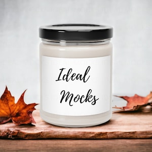 Peut inclure: Une bougie en pot de verre transparent avec un couvercle noir et une étiquette blanche. L'étiquette indique "Ideal Mocks" en écriture noire. La bougie est sur une surface en bois avec des feuilles d'automne.