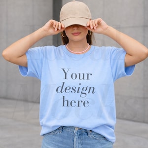 May include: A light blue t-shirt with the text "Your design here" in a dark gray font. The person is wearing a tan baseball cap and blue jeans. The person is also wearing a pink pearl necklace and gold hoop earrings.