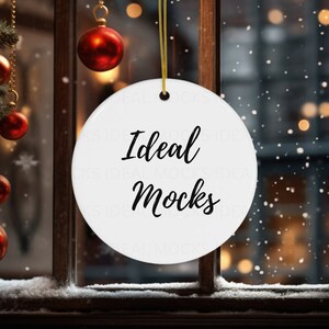 May include: A round white ceramic ornament hanging by a gold string. The ornament features the black text Ideal Mocks printed in the center. The background shows a window with snow, pine branches, and red ornaments, creating a festive holiday scene.