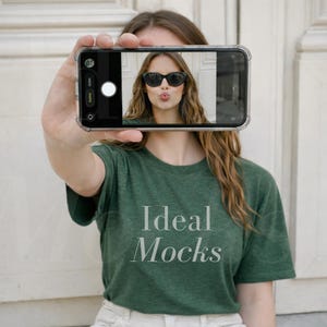 May include: A green t-shirt with the text "Ideal Mocks" in white. The image on the phone screen shows a person wearing sunglasses. The phone is held up in front of the person.