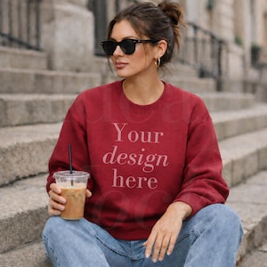 May include: A maroon sweatshirt with the text "Your design here" in a light pink font. The person is wearing sunglasses and holding a clear plastic cup with a black straw. The person is wearing blue jeans.