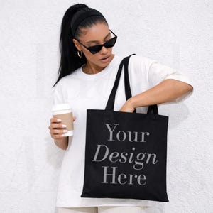 以下が含まれることがあります： 「Your Design Here」の文字がセリフ体でプリントされた黒いトートバッグ。長い持ち手があり、白いTシャツとサングラスをかけた人が持っています。